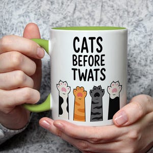 May include: White ceramic mug with a green handle and interior. The mug features the text "CATS BEFORE TWATS" in black, along with illustrations of four different cat paws. The paws are in various colors, including black, orange, and gray.