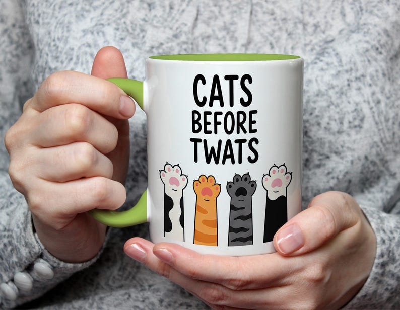 May include: White ceramic mug with a green handle and interior. The mug features the text "CATS BEFORE TWATS" in black, along with illustrations of four different cat paws. The paws are in various colors, including black, orange, and gray.