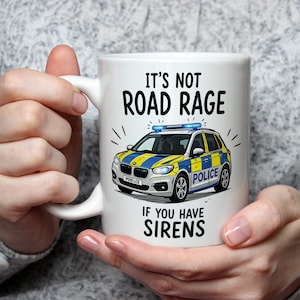 Può includere: Tazza in ceramica bianca con un'illustrazione di un'auto della polizia con dettagli blu e gialli. La tazza presenta il testo "It's not road rage if you have sirens". La tazza è tenuta da una persona.