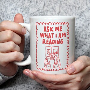 Op de afbeelding: Witte keramische mok met een rood ontwerp met de tekst "ASK ME WHAT I AM READING". Het ontwerp bevat een tekening van een persoon die een boek leest. De mok wordt in de handen van een persoon gehouden.