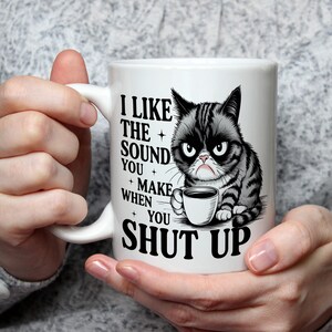 Könnte beinhalten: Weiße Keramik-Tasse mit einer Illustration einer grummeligen Katze und dem Text "I LIKE THE SOUND YOU MAKE WHEN YOU SHUT UP." Die Katze ist schwarz und grau und hält eine Kaffeetasse. Die Tasse hat einen gebogenen Henkel.