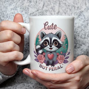 Puede incluir: Taza de cerámica blanca con un diseño de mapache de dibujos animados, un corazón y rosas. La taza tiene el texto "Cute But Feral". La taza está sostenida por una persona.