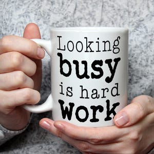 May include: White ceramic mug with the text "Looking busy is hard work" in black lettering. The mug is held by a person wearing a gray sweater. The mug has a curved handle.
