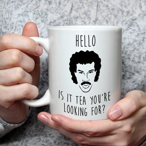 May include: White ceramic mug featuring a black illustration of a man with a mustache and curly hair. The mug has the text "HELLO IS IT TEA YOU'RE LOOKING FOR?" in black. The mug is held by a person.