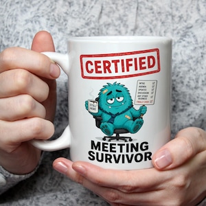 May include: A white ceramic mug featuring a graphic of a teal monster sitting in an office chair, wearing a tie, and holding a mug labelled STILL HERE. Above the monster is the text CERTIFIED and below is MEETING SURVIVOR with a checklist.