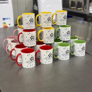 Pode incluir: Uma pilha de canecas de cerâmica branca com um logotipo preto em forma de pata que diz "The Dog Shop". As canecas têm bordas de cores diferentes, incluindo vermelho, amarelo e verde.