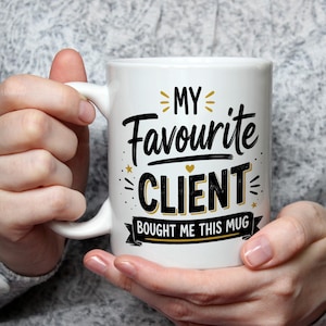 以下が含まれることがあります： MY FAVOURITE CLIENT BOUGHT ME THIS MUGという黒と金の文字が書かれた白いセラミック製のマグカップ。デザインには小さな金の星と文字を囲む装飾的な線が含まれており、最後のフレーズは黒いバナーの上に配置されています。
