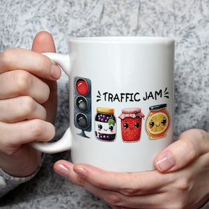 May include: White ceramic mug featuring a red, yellow, and green traffic light graphic and the text "TRAFFIC JAM" with three cartoon jars of jam with faces. The jars are filled with blueberry, strawberry, and orange jam.