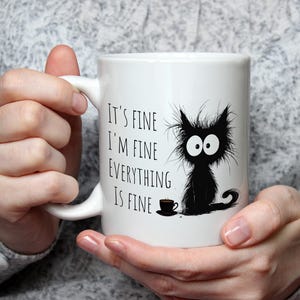 May include: White ceramic mug with a black cartoon cat and the text "It's fine, I'm fine, everything is fine." A small coffee cup is next to the cat. The mug is held by a person.