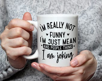 May include: White ceramic mug with the text "I'M REALLY NOT FUNNY I'M JUST MEAN AND PEOPLE THINK I am joking" in black script. The mug has a curved handle and is held by a person.