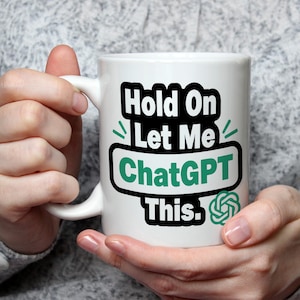 May include: White ceramic mug with the text "Hold On Let Me ChatGPT This." in black and green. The mug also features the ChatGPT logo. The mug is held by a person.