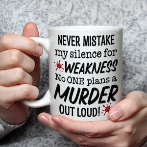 以下が含まれることがあります： 「NEVER MISTAKE my silence for WEAKNESS No ONE plans a MURDER OUT LOUD!」の黒い文字が書かれた白いセラミックマグ。赤い飛沫がテキストを強調しています。マグカップは湾曲したハンドルがあり、人が持っています。