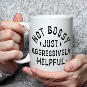 May include: White ceramic mug with the text "NOT BOSSY JUST AGGRESSIVELY -HELPFUL-" in black. The mug has a curved handle and is held by a person wearing a gray sweater.