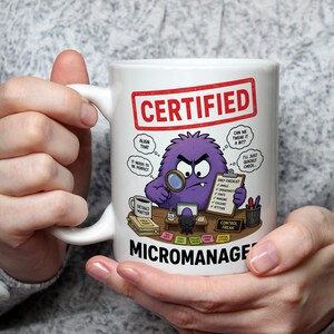 May include: A white ceramic mug featuring a graphic of a purple monster at a desk with the text CERTIFIED MICROMANAGER. The illustration includes a magnifying glass, a daily checklist, sticky notes, and a sign that reads CONTROL FREAK.