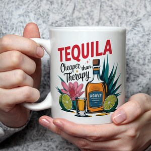 以下が含まれることがあります： 「TEQUILA Cheaper than Therapy」の文字が赤と黒でプリントされた白いセラミックマグ。テキーラボトル、ショットグラス、ライムスライス、花の装飾が施されています。マグカップは人が持っています。