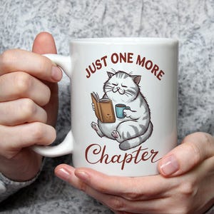 Könnte beinhalten: Weiße Keramiktasse mit einer Cartoon-Katze, die ein Buch liest und eine blaue Tasse hält. Auf der Tasse steht in brauner Schrift "JUST ONE MORE CHAPTER". Die Katze ist grau und weiß.