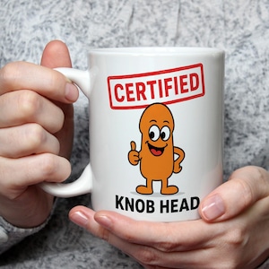 May include: A white ceramic mug featuring a cartoon character that is orange, oval-shaped, and smiling with a thumbs-up gesture. Above the character is a red rectangular stamp with the text CERTIFIED, and below is the text KNOB HEAD.