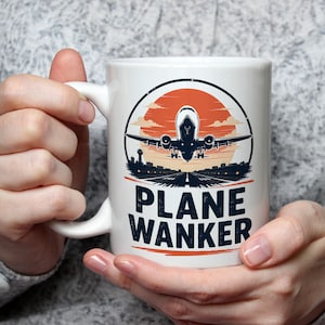 May include: A white ceramic mug featuring a graphic illustration of an airplane taking off from a runway against a large orange sun. Below the illustration, the text PLANE WANKER is printed in bold, dark blue capital letters on the front surface.
