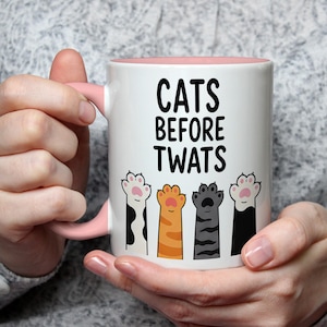 May include: White ceramic mug with a pink interior and handle. The mug features the text "CATS BEFORE TWATS" in black, with illustrations of colorful cat paws below the text. The mug is held by a person.