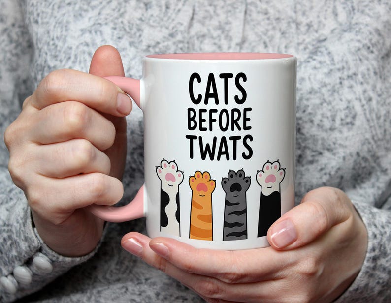 May include: White ceramic mug with a pink interior and handle. The mug features the text "CATS BEFORE TWATS" in black, with illustrations of colorful cat paws below the text. The mug is held by a person.
