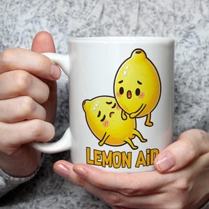 Può includere: Tazza in ceramica bianca con un'immagine di due limoni di cartone animato, uno sopra l'altro. Il testo "Lemon Aid" è stampato sotto i limoni.