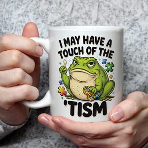 Puede incluir: Taza de cerámica blanca con una ilustración de una rana verde y el texto "I MAY HAVE A TOUCH OF THE 'TISM". La rana sostiene un fidget spinner y apunta hacia arriba. Piezas de rompecabezas coloridas rodean la rana.
