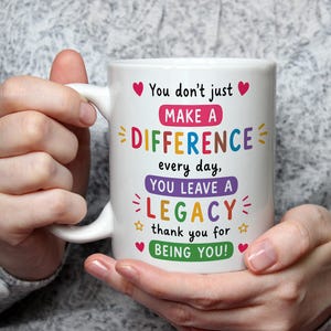 May include: A white ceramic mug featuring colorful text that reads: You don't just MAKE A DIFFERENCE every day, YOU LEAVE A LEGACY thank you for BEING YOU! The design includes small red hearts, yellow stars, and decorative dashes in various colors.