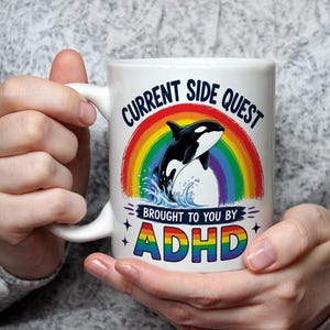 May include: White ceramic mug featuring a colorful design with a rainbow, a black and white orca, and the text "CURRENT SIDE QUEST BROUGHT TO YOU BY ADHD". The mug has a curved handle.