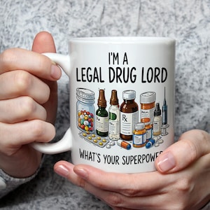 May include: White ceramic mug featuring the text "I'M A LEGAL DRUG LORD WHAT'S YOUR SUPERPOWER?" and illustrations of pills, medicine bottles, and a syringe. The mug is held by a person wearing a grey jumper.