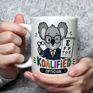 May include: A white ceramic mug featuring a cartoon koala wearing glasses and a suit, holding a handheld eye exam tool next to an eye chart. The text KOALIFIED OPTICIAN is printed in colourful letters below the koala illustration.