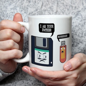 Könnte beinhalten: Weiße Keramik-Tasse mit einem humorvollen Design, das eine Diskette und einen USB-Stick mit Sprechblasen zeigt. Die Diskette sagt "I AM YOUR FATHER" und der USB-Stick antwortet "NOPEEEEE!".