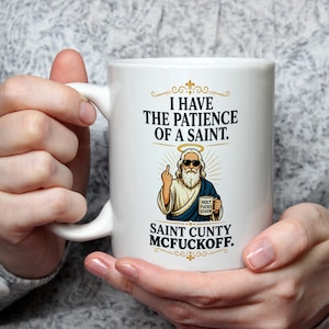 May include: White ceramic mug with a humorous graphic. The design features an illustration of a saint giving the middle finger, with the text "I HAVE THE PATIENCE OF A SAINT. SAINT CUNTY MCFUCKOFF."