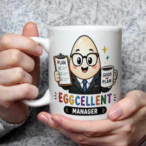 Può includere: Una tazza in ceramica bianca con un personaggio a forma di uovo in stile cartoon che indossa occhiali e un abito. L'uovo tiene in mano una cartellina con scritto PLAN e una tazza con scritto GOOD PLAN. Sotto, EGGCELLENT MANAGER.