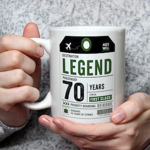 May include: A white ceramic mug featuring a graphic design styled like an airline luggage tag. The text reads DESTINATION LEGEND, PASSENGER 70 YEARS, STATUS FIRST CLASS, PRIORITY BOARDING, and BAGGAGE 70 YEARS OF STORIES, with an airplane icon.