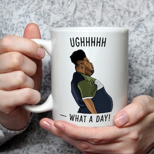 Op de afbeelding: Witte keramische mok met een cartoon illustratie van een man en de tekst "UGHHHHHH - WHAT A DAY!". De man draagt een blauw, groen en wit shirt. De mok wordt door een persoon vastgehouden.