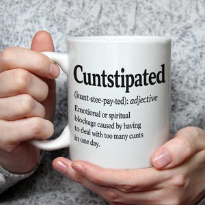 May include: A white ceramic mug featuring black printed text that defines the word Cuntstipated as an adjective, describing an emotional or spiritual blockage caused by having to deal with too many cunts in one day, including phonetic pronunciation.