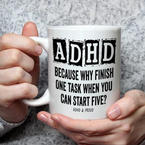May include: White ceramic mug featuring the text "ADHD BECAUSE WHY FINISH ONE TASK WHEN YOU CAN START FIVE?" in black. The mug also has the text "ADHD & PROUD". The mug is held by a person.