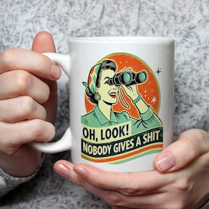 Może przedstawiać: Biały ceramiczny kubek z retro ilustracją kobiety patrzącej przez lornetkę. Kubek ma napis "OH, LOOK! NOBODY GIVES A SHIT" na banerze pod ilustracją.