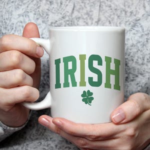 May include: White ceramic mug with the word "IRISH" in green block letters and a green four-leaf clover. The mug is held by a person wearing a grey sweater.
