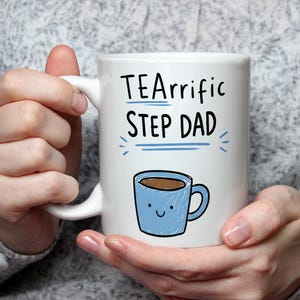 Puede incluir: Una taza de cerámica blanca con el texto negro "TEArrific STEP DAD" y líneas decorativas azules debajo. Debajo del texto hay una pequeña ilustración de una taza azul claro con una cara sonriente y líquido marrón en su interior.