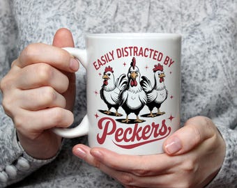 May include: White ceramic mug featuring a humorous design with three cartoon chickens. The text "EASILY DISTRACTED BY Peckers" is printed in red. The mug is held by a person.