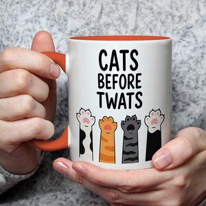 May include: White ceramic mug with an orange interior and handle. The mug features the text "CATS BEFORE TWATS" in black, with illustrations of cat paws below. The paws are in various colors and patterns.