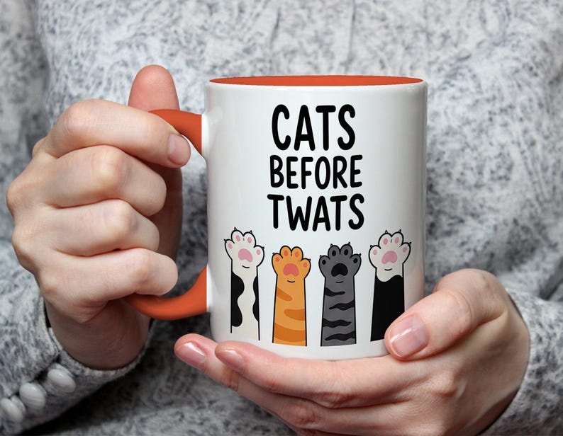 May include: White ceramic mug with an orange interior and handle. The mug features the text "CATS BEFORE TWATS" in black, with illustrations of cat paws below. The paws are in various colors and patterns.