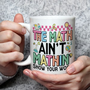 May include: White ceramic mug with the text "THE MATH AIN'T MATHIN' SHOW YOUR WORK" in colourful, playful lettering. The mug features illustrations of school-related items like a book, scissors, and a globe. The mug is held by a person.