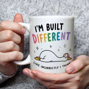 Può includere: Tazza in ceramica bianca con la scritta "I'M BUILT DIFFERENT" in nero e colori arcobaleno. Un'anatra dei cartoni animati con un cappello è sdraiata sotto il testo. La frase "LIKE INCORRECTLY I THINK" è scritta sotto.