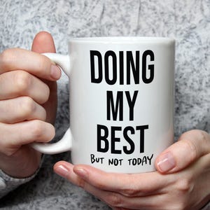 May include: White ceramic mug with the text "DOING MY BEST BUT NOT TODAY" in black, held by a person. The mug has a curved handle and a cylindrical shape. The background is a grey, textured fabric.