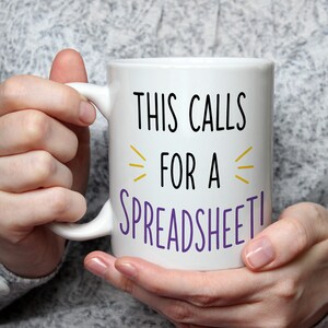 Könnte beinhalten: Ein weißer Keramikbecher mit dem schwarzen Text THIS CALLS, gelben Zierlinien und dem violetten Text FOR A SPREADSHEET! Der Becher hat einen Standardhenkel und eine glatte Oberfläche, die eine klare Sicht auf den Aufdruck auf der Vorderseite bietet.