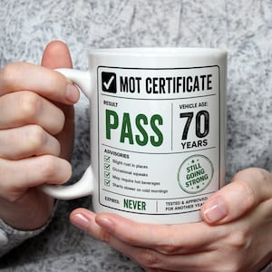 May include: A white ceramic mug featuring a graphic design styled as an MOT certificate. The text reads MOT CERTIFICATE, RESULT PASS, VEHICLE AGE 70 YEARS, and ADVISORIES including slight rust, occasional squeaks, and a stamp stating STILL GOING STRONG.