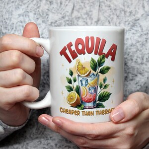 以下が含まれることがあります： 赤いブロック体の「TEQUILA」の文字が入った白いセラミックマグ。マグには、氷、レモンスライス、緑の葉が入ったカクテルのカラフルなイラストが描かれています。「cheaper than therapy」の文字が下にあります。