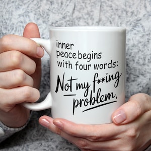 以下が含まれることがあります： 「inner peace begins with four words: Not my f**king problem」の黒い文字が書かれた白いセラミックマグ。グレーのセーターを着た人がマグカップを持っています。